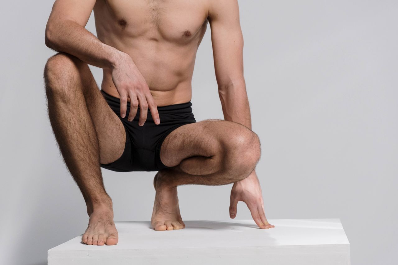 Close-up of a shirtless man squatting, wearing black underwear on a white platform.
